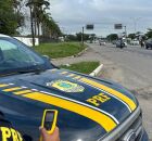 Agente ao lado de viatura da PRF em rodovia federal.