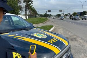 Agente ao lado de viatura da PRF em rodovia federal.