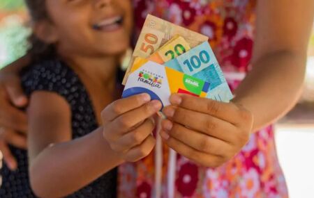 Cart&atilde;o e dinheiro do Bolsa Fam&iacute;lia sendo segurados por m&atilde;e e crian&ccedil;a.