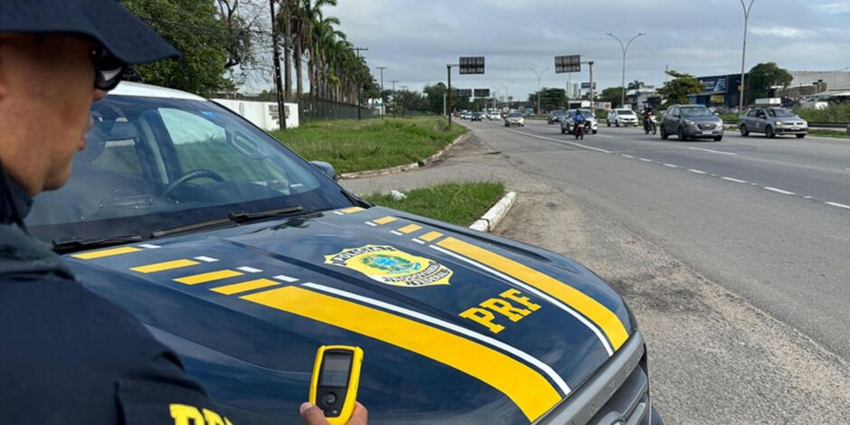 Agente ao lado de viatura da PRF em rodovia federal.