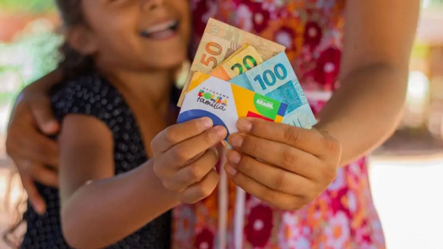 Cartão e dinheiro do Bolsa Família sendo segurados por mãe e criança.